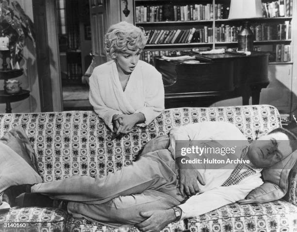 Richard, played by Tom Ewell lies on a sofa hugging his stomach whle a sympathetic Marily Monroe playing 'The Girl' leans over him. A scene from 'The...