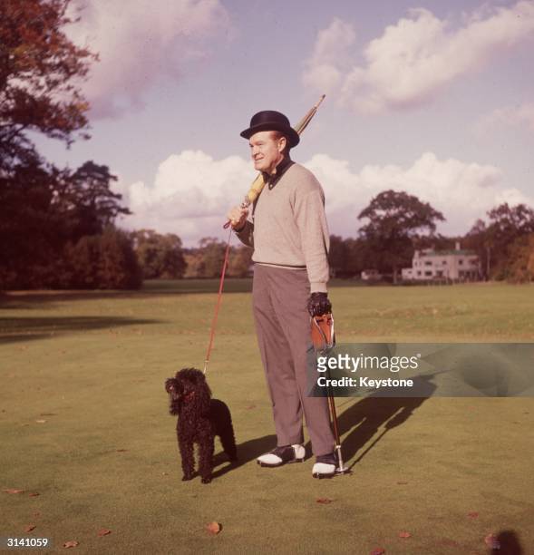 British-born American comedian and actor Bob Hope out walking on an English golf course with an umbrella, a shooting stick and a black poodle.
