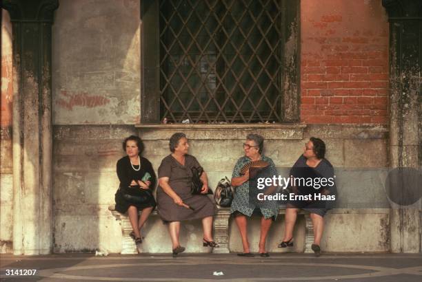 Elderly Venetian women of goodly proportions sit on a stone bench gossiping and fanning themselves in the shade of an arched passageway in the Piazza...