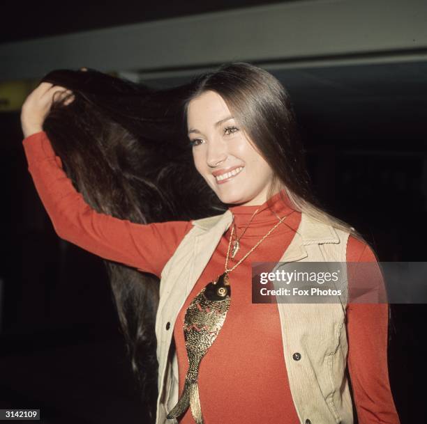 English actress Jane Seymour shows off her famously long dark hair upon arrival at London Airport. Around her neck is a large fish pendant.