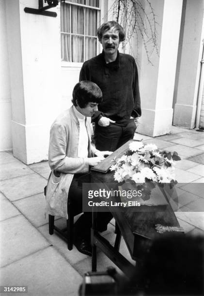 Irish actor Richard Harris stands beside songwriter Jim Webb who is playing a keyboard which is decorated with a vase of flowers.