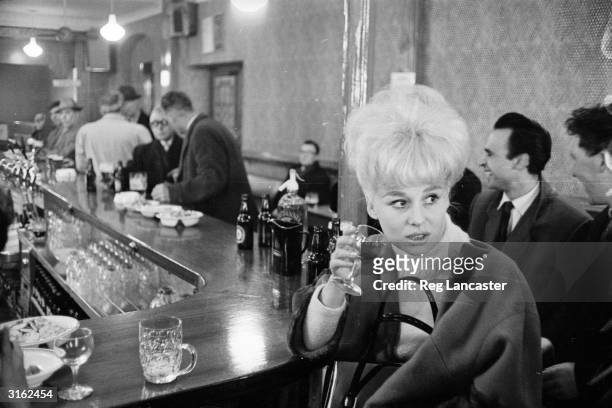 English actress Barbara Windsor enjoying a drink at the bar of a pub.