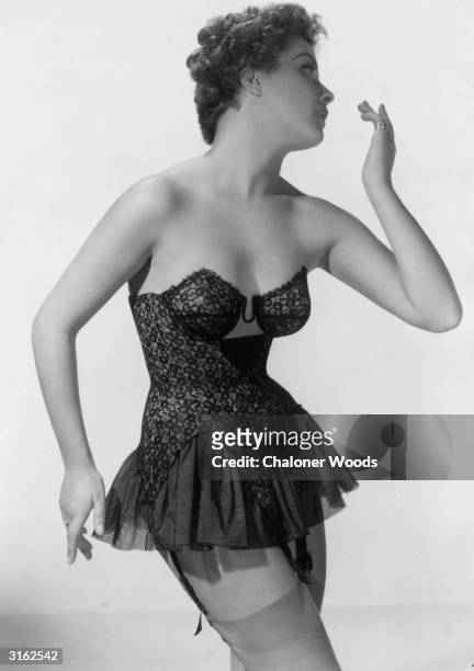 Woman modelling a black lace corset with underwired cups, a short frilled skirt and suspenders.