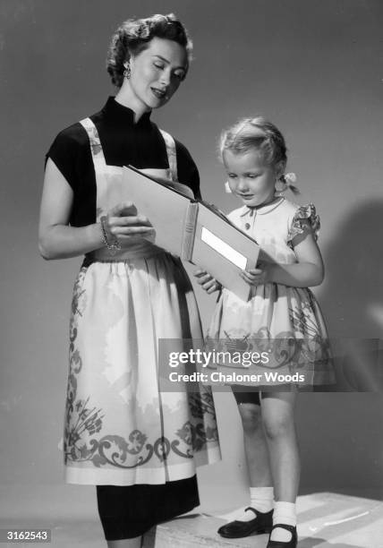 Woman helps her little girl read from the Good Housekeeping Picture Cookery book. The mother is wearing a souvenir apron with a London theme, while...