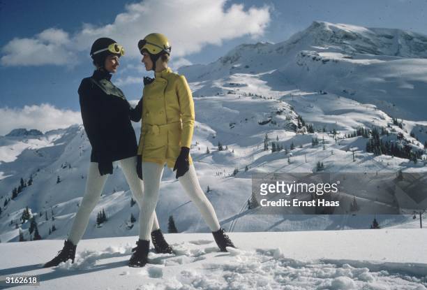 Two women pose on a mountain in the Alps, wearing ski helmets, coloured jackets and white leggings.