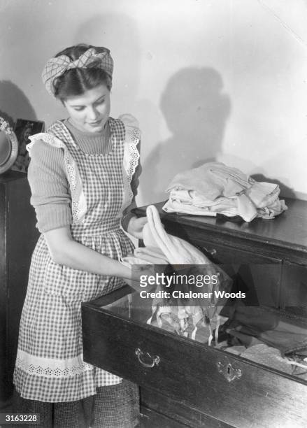Housewife wearing a gingham and lace apron places washed and ironed clothing into a chest of drawers.