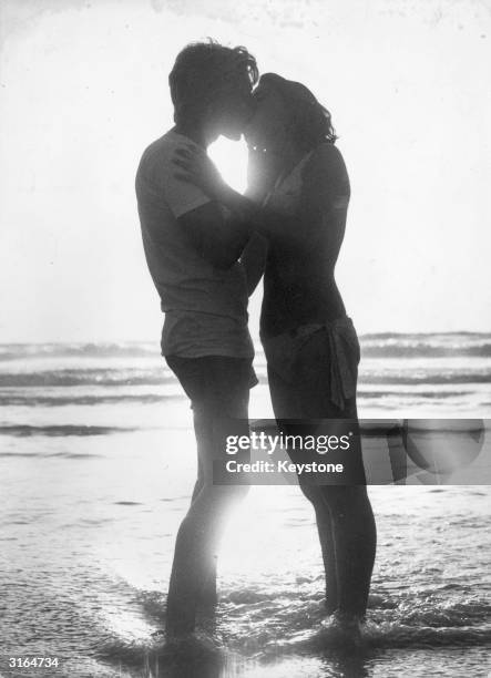 Young lovers stand in the surf as they kiss at sunset.