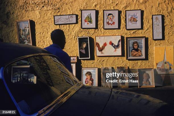 Man leans against a car as he looks at a display of paintings hung on a stuccoed wall in New York village.
