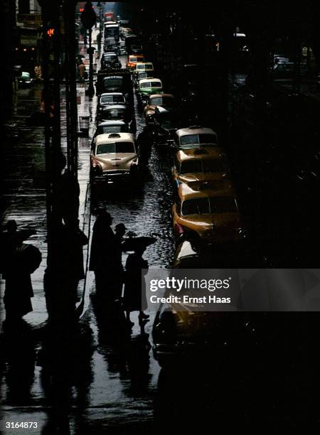 At the traffic lights pedestrians are crossing a street full of cars and yellow cabs on a dark, wet day in New York.