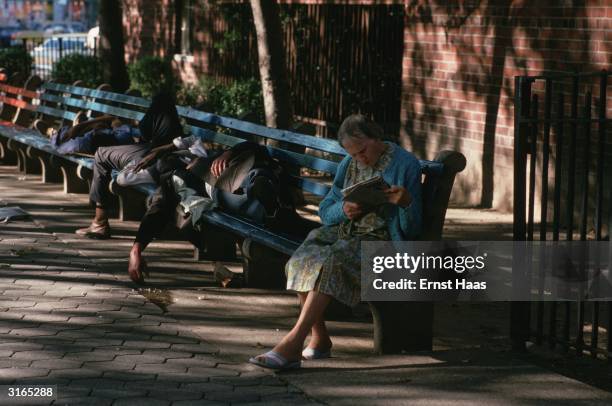 In a New York street down and outs sprawl asleep on a bench at the end of which an old woman sits reading a magazine.