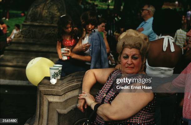 Large and querulous looking New Yorker wearing a large wig is sitting in the park on a summer's day.