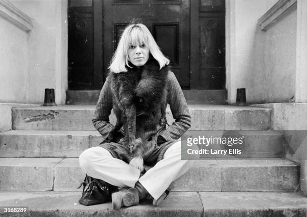 Italian-born German actress Anita Pallenberg sits cross-legged on a flight of stone steps with her hands in her pockets.
