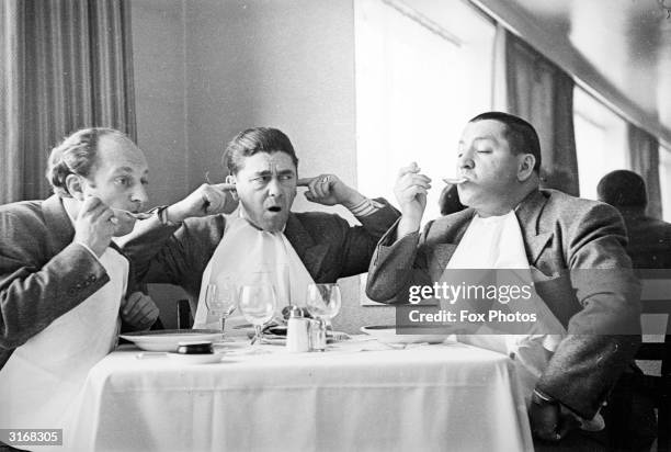 American comedian Moe Howard sticking his fingers in his ears while his fellow comedians, Larry Fine and Curly Howard noisily eat soup. The trio...