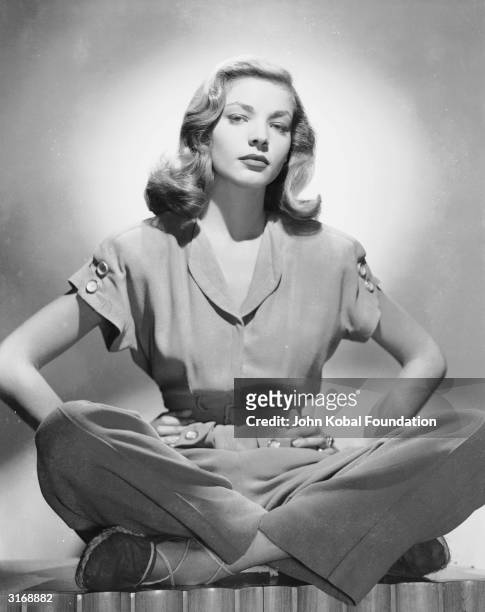 American screen star Lauren Bacall sitting cross-legged in a trouser suit, with her hands on her hips.