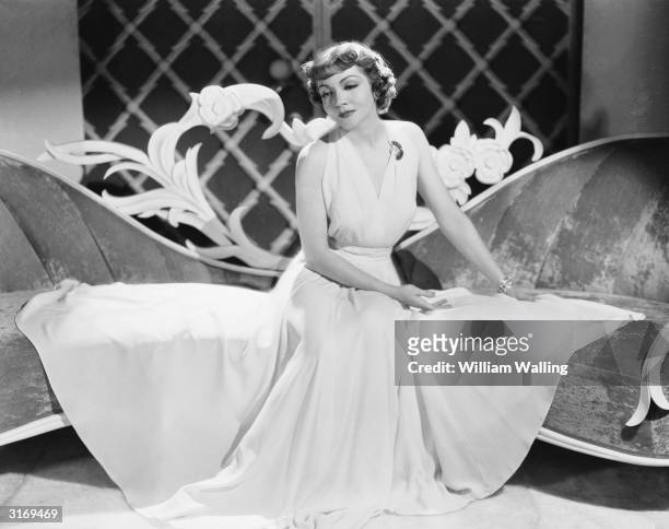 French born actress Claudette Colbert sits on an unusually-shaped sofa with her skirt fanning out around her.