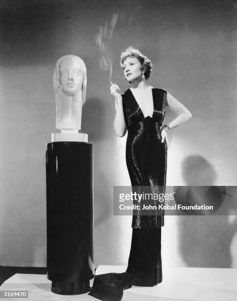 French born actress Claudette Colbert stands next to a sculpture smoking a cigarette.