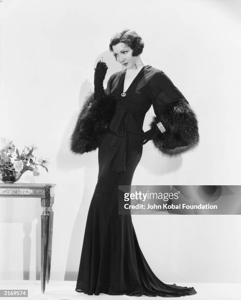 French born actress Claudette Colbert wearing a full length gown with fur cuffs and long gloves for her role as Felicia Hammond in 'The Phantom...