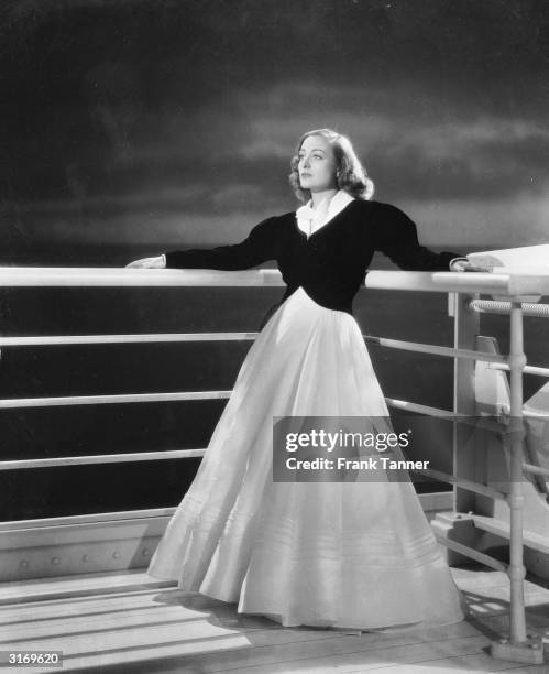 Leaning against a ship's rails, film star Joan Crawford in a full length full skirted evening dress worn with a long sleeved, waist length, jacket....