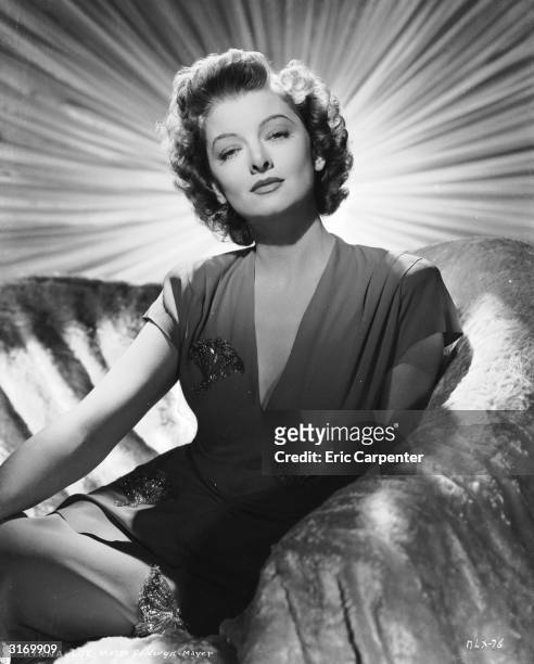 American actress Myrna Loy sitting on a covered sofa with a curtain with a bright sunshine design behind her.