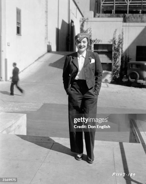 Casual Marlene Dietrich dressed in slacks, jacket and hat smiles in the sunshine with her hands in her pockets.