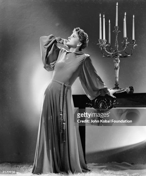 British-born actress Joan Fontaine strikes a theatrical pose in a floor-length belted gown with full sleeves gathered at the wrist. An ornate...