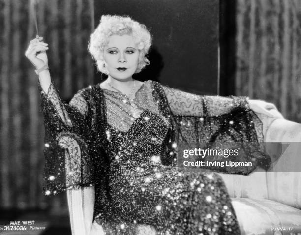 Dressed in a sequinned gown, Mae West lounges with a cigarette in her first screen role as Maudie in 'Night After Night', directed by Archie Mayo.