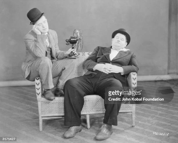 Sleeping Stan Laurel perches on the arm of a wicker sofa, pointing a fan at Oliver Hardy .