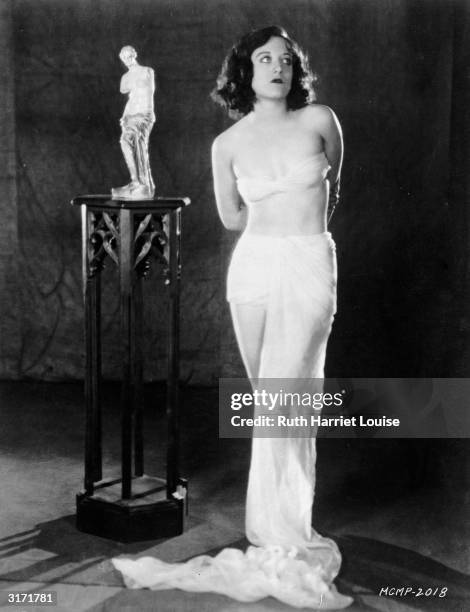 With her arms held behind her back and in bra and draperies, film star Joan Crawford poses beside a small statue of the Venus de Milo.