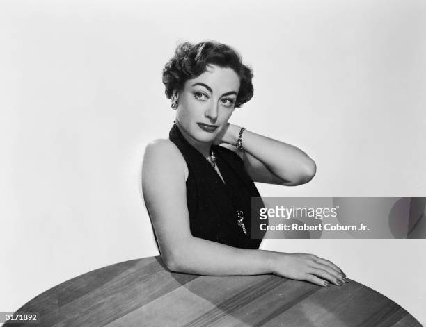 American film actress Joan Crawford leaning on a table in a promotional picture for her latest film 'Harriet Craig'.