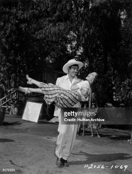 Full-length image of Mexican-born actor Anthony Quinn carrying American actor Dorothy Lamour down the South Sea Island street where director Victor...