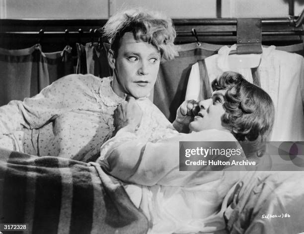 American actor Tony Curtis grabs American actor Jack Lemmon by the collar of his nightgown and makes a fist as they lie in a bed together, dressed in...