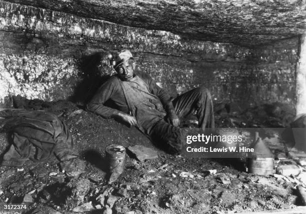 Coal miner leans back on his elbow, preparing to eat his lunch. There is a metal lunchpail and pitcher in the dirt. He has his helmet and overalls...