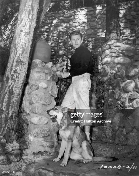 American actor Kirk Douglas, standing beside a stone wall and well, prepares to take his German shepherd, Banshee, for a walk during a break in the...