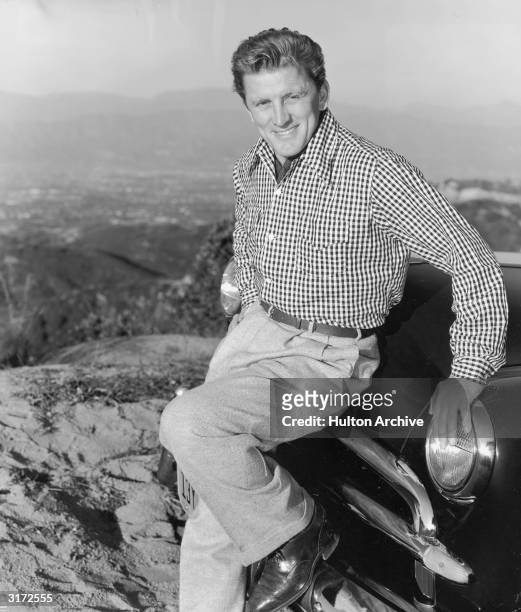 Portrait of American actor Kirk Douglas leaning against the hood of a car and resting his foot on the bumper. Douglas is posing on top of a mountain...