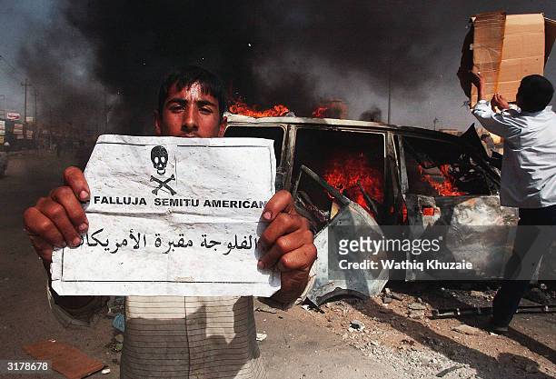 An Iraqi boy holds a sign that reads "Fallujah is the cemetary of the Americans" as a sport utility vehicle burns behind him following an attack on...