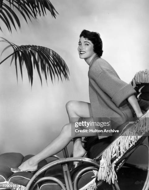 Portrait of American actor Julie Adams posing on a lounge chair in a cardigan and metallic mules with palm leaves in the background.