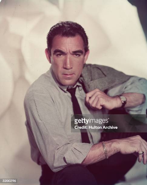 Studio portrait of Mexican-born actor Anthony Quinn sitting and holding a sports coat over his shoulder. He is wearing a shirt, tie and silver...