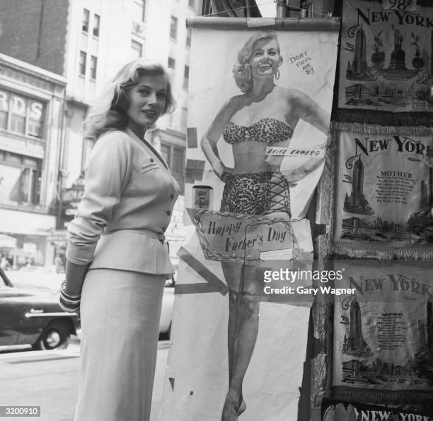 Swedish-born actor Anita Ekberg, wearing a skirt suit, poses next to a full-length poster of herself wearing a two-piece leopard print swimsuit,...