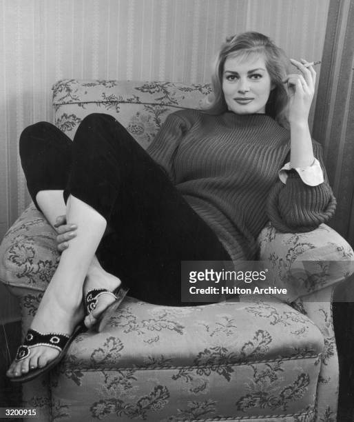 Full-length image of Swedish-born actor Anita Ekberg, wearing a sweater and Capri pants, sitting with her feet up on an armchair and smoking a...