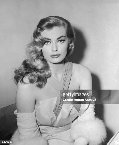 Seated portrait of Swedish-born actor Anita Ekberg, wearing a dress with crisscrossed straps and a cardigan with fur trim, 1950s. Ekberg looks as she...