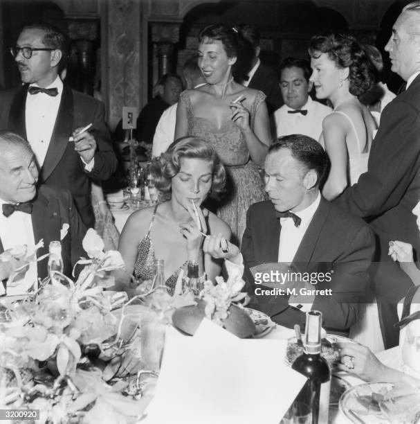 American actor and singer Frank Sinatra sits at a table lighting the cigarette of American actor Lauren Bacall, at a party following the premiere of...