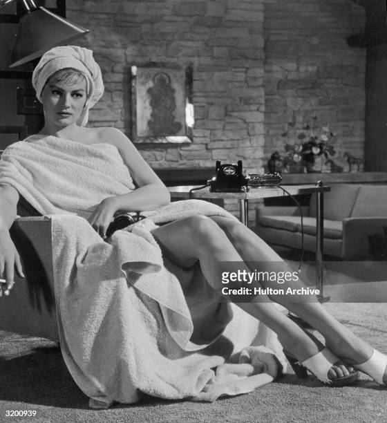 Portrait of Swedish-born actor Anita Ekberg seated with towels wrapped around her toga-style, smoking a cigarette, with a telephone receiver in her...