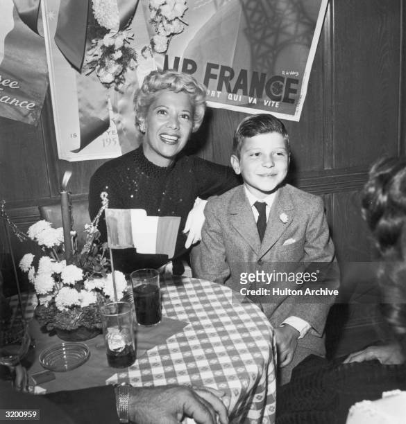 American singer Dinah Shore, wearing a dark sequined gown, smiles while sitting at a table with young Frank Sinatra Jr., who wears a suit.