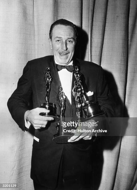 American cartoonist and producer Walt Disney laughs while struggling to hold four Oscar statuettes simultaneously, backstage at the Academy Awards,...