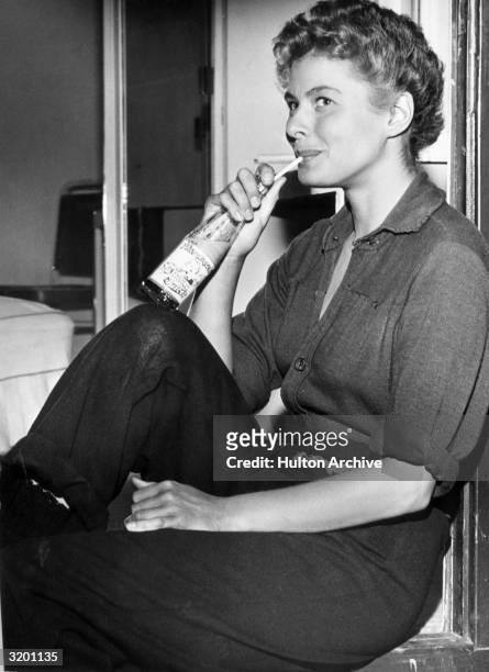 Swedish-born actor Ingrid Bergman sits on a window ledge, sipping juice from a glass bottle with a straw, on the set of director Sam Wood's film,...