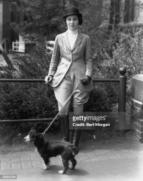Full-length view of Mrs. John V. Bouvier III, wearing a riding habit, smoking a cigarette while holding her spaniel 'Bonnett' on a leash at a...