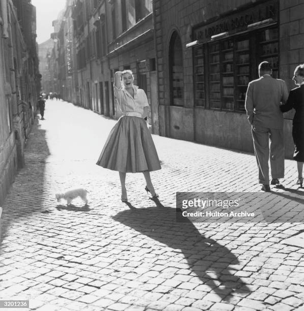 Full-length portrait of Swedish-born actor Anita Ekberg, wearing a full skirt with a striped blouse, laughing while posing next to a toy poodle on a...