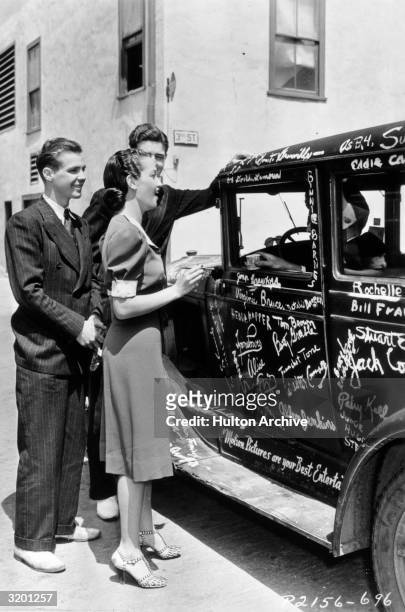Full-length view of American actor Dorothy Lamour, standing openmouthed with a paintbrush, before signing a dark car covered with celebrity...
