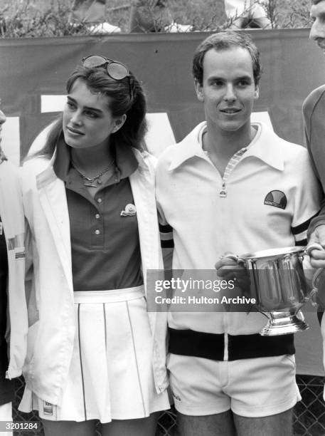 American model and actor Brooke Shields stands with Prince Albert of Monaco. The Prince holds a sports trophy cup. Both are wearing tennis outfits.