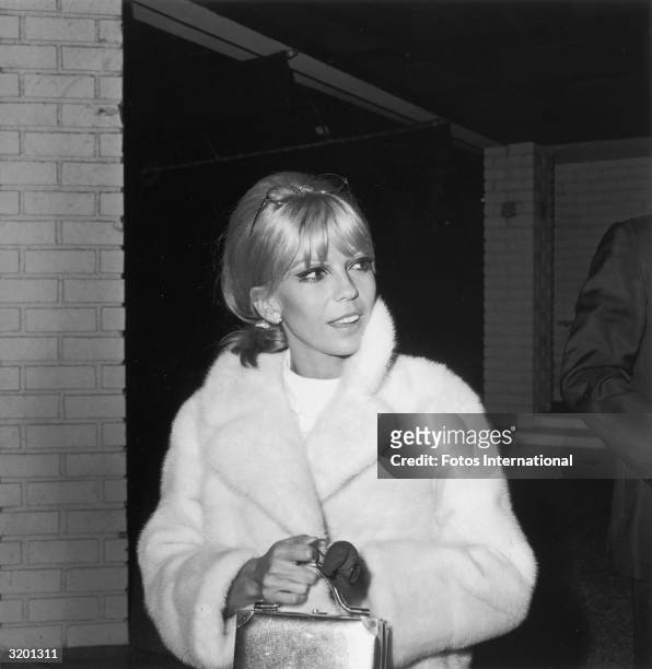 American singer Nancy Sinatra holds a box bag while wearing a light-colored fur coat. Sinatra has blonde hair and wears dramatic eye makeup,...